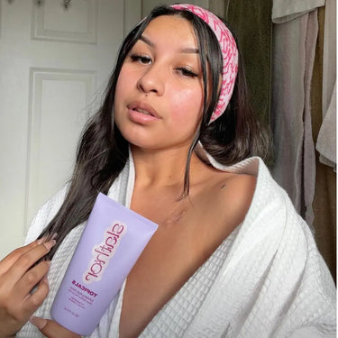 Face brightening treatment featured by a woman in a white robe, holding a pastel purple skincare product in a bathroom setting
